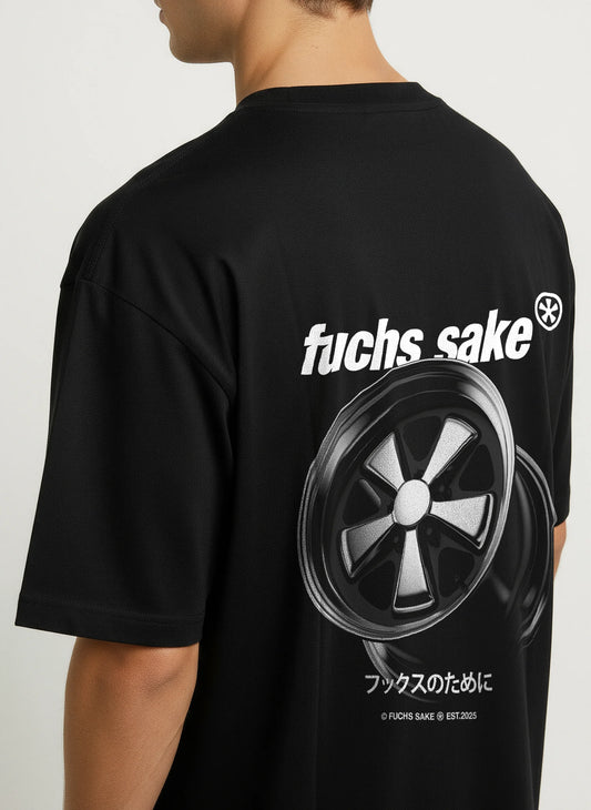 Side view of man wearing black ‘fuchs sake’ T-shirt with bold white logo and silver Fuchs wheel graphic on back — classic Porsche-inspired streetwear for car enthusiasts.