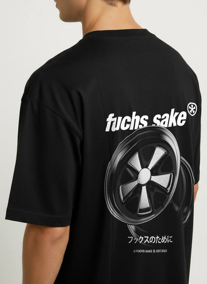 Side view of man wearing black ‘fuchs sake’ T-shirt with bold white logo and silver Fuchs wheel graphic on back — classic Porsche-inspired streetwear for car enthusiasts.