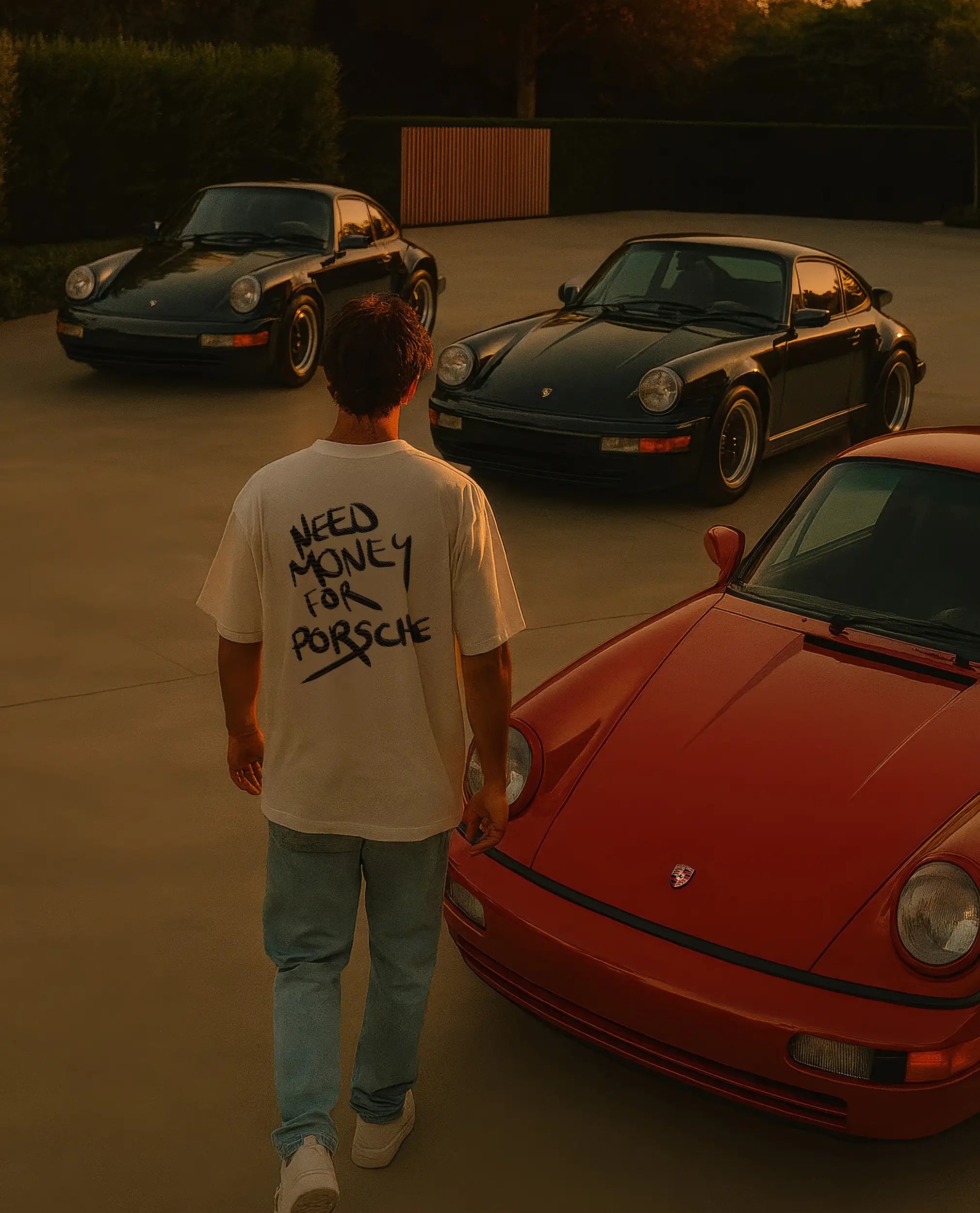 Man standing in front of a red Porsche with two other Porsches in the background and wearing a white tshirt reading "Need Money For Porsche"