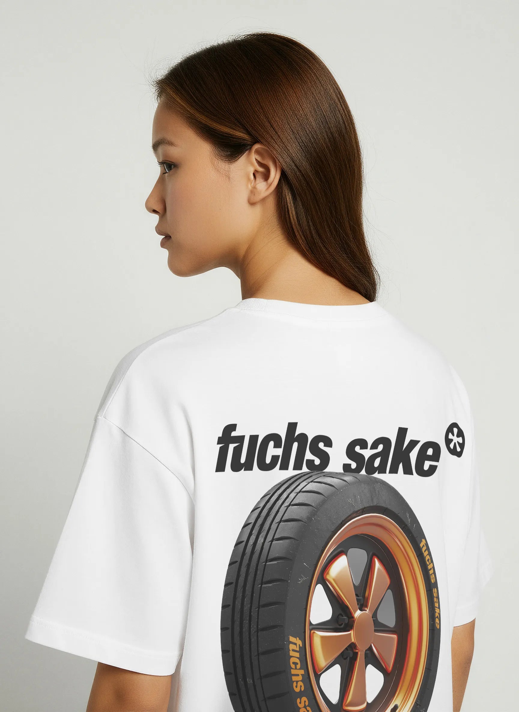 Side view of woman wearing white T-shirt with large rear print featuring a bronze Porsche Fuchs wheel and bold ‘fuchs sake’ text — car enthusiast apparel inspired by classic Porsche design.